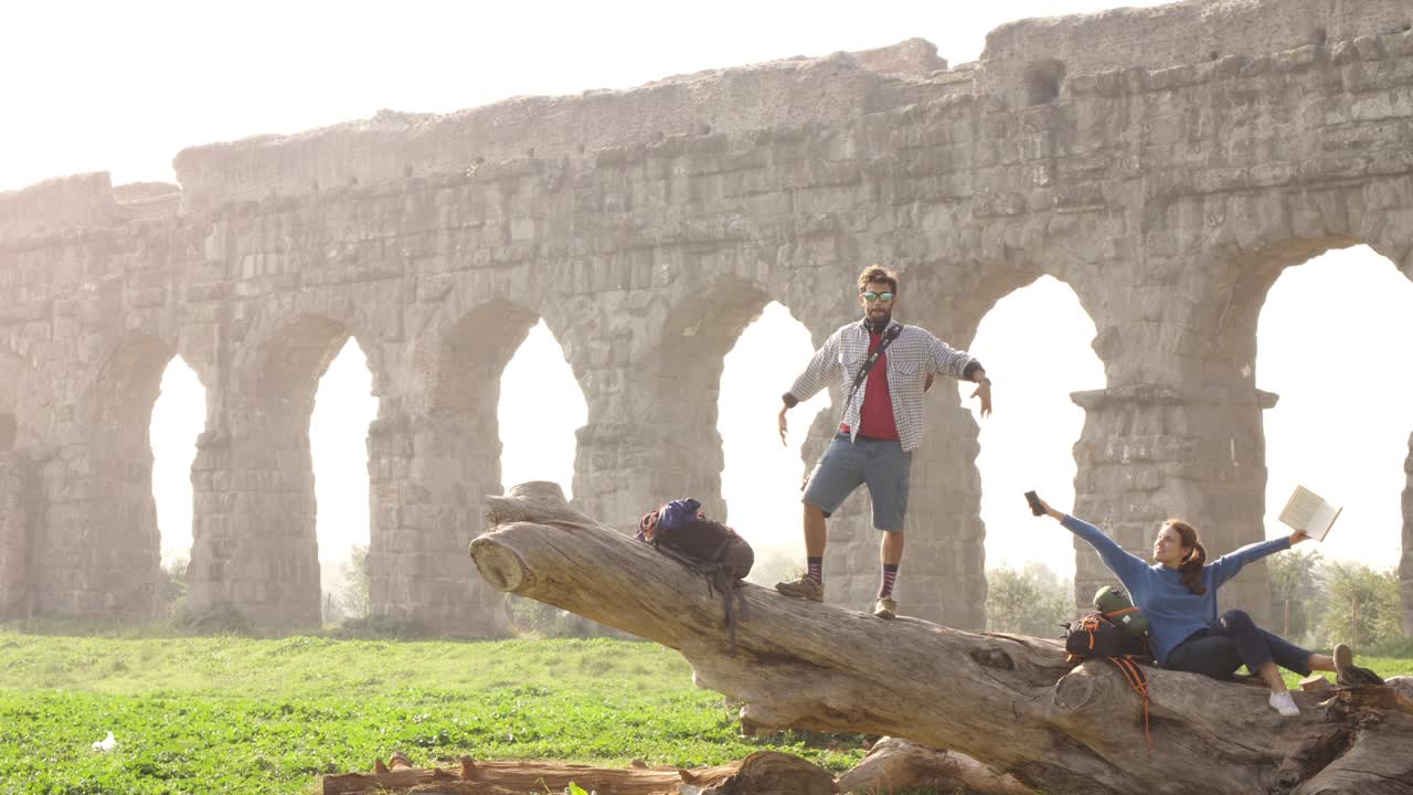 счастливая молодая пара туристов с гитарой на бревне позирует для фотографии перед древнеримским акведуком руины в романтическом парке parco degli aquedotti парк в риме на туманном восходе солнца замедленное движение