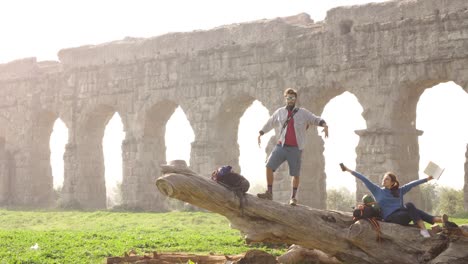 happy young couple backpackers tourists with guitar on a log trunk posing for picture in front of ancient roman aqueduct ruins in romantic parco degli acquedotti park in rome at misty sunrise slow motion