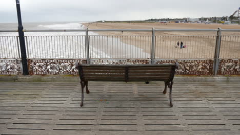 An empty bench on n abandoned pier overlooks a sandy beach and the distant sea under an overcast sky
