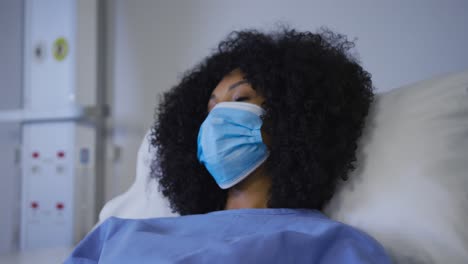 Portrait-of-african-american-female-patient-wearing-face-mask-lying-on-hospital-bed