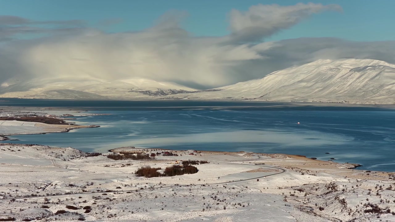 Riprese aeree catturano Eyjafjörður, Islanda, mentre le acque blu intenso del fiordo riflettono un cielo nuvoloso, circondate da neve immacolata, strade tortuose, dolci colline e un drammatico paesaggio costiero invernale.