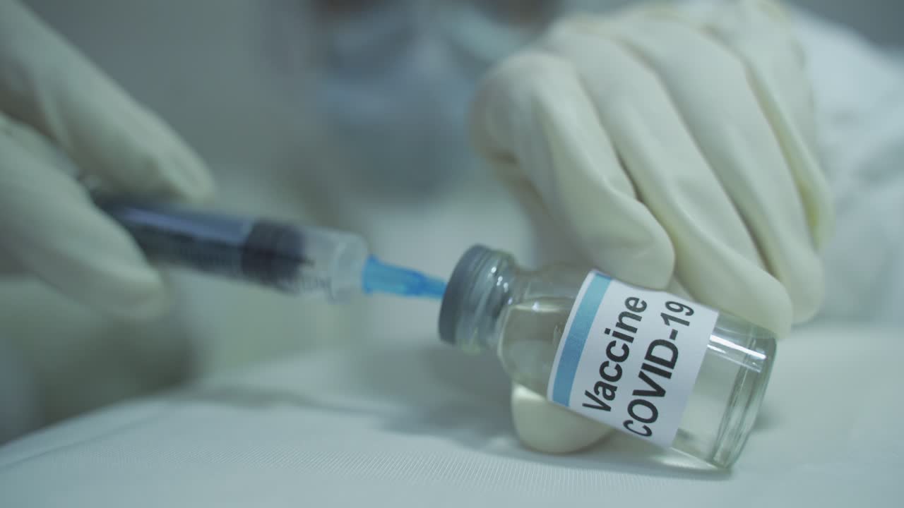 human wearing protective PPE kit carefully drawing out covid 19 vaccine from vial and filling into syringe in horizontal position