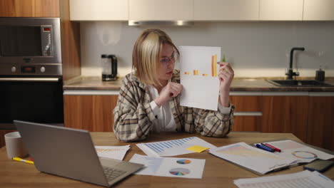 a woman with glasses at home shows a graph to a laptop camera while sitting at home in the kitchen. home office remote work. video call and conference with graphs and tables.