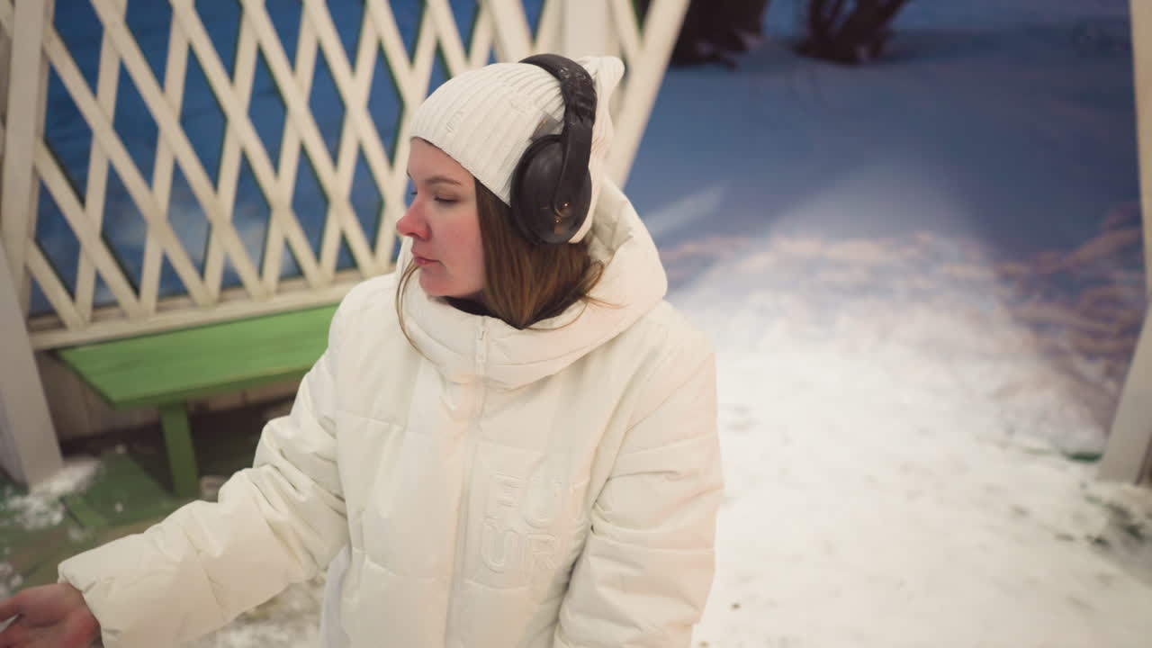 Fair lady wearing white puffer coat and headphones dances inside light decorated summer house in snowy environment opening and closing eyes expressively under warm glow of overhead bulbs