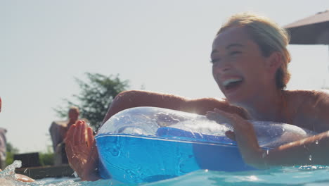 Couple-With-Man-Splashing-Woman-Floating-On-Airbed-In-Swimming-Pool-On-Summer-Holiday