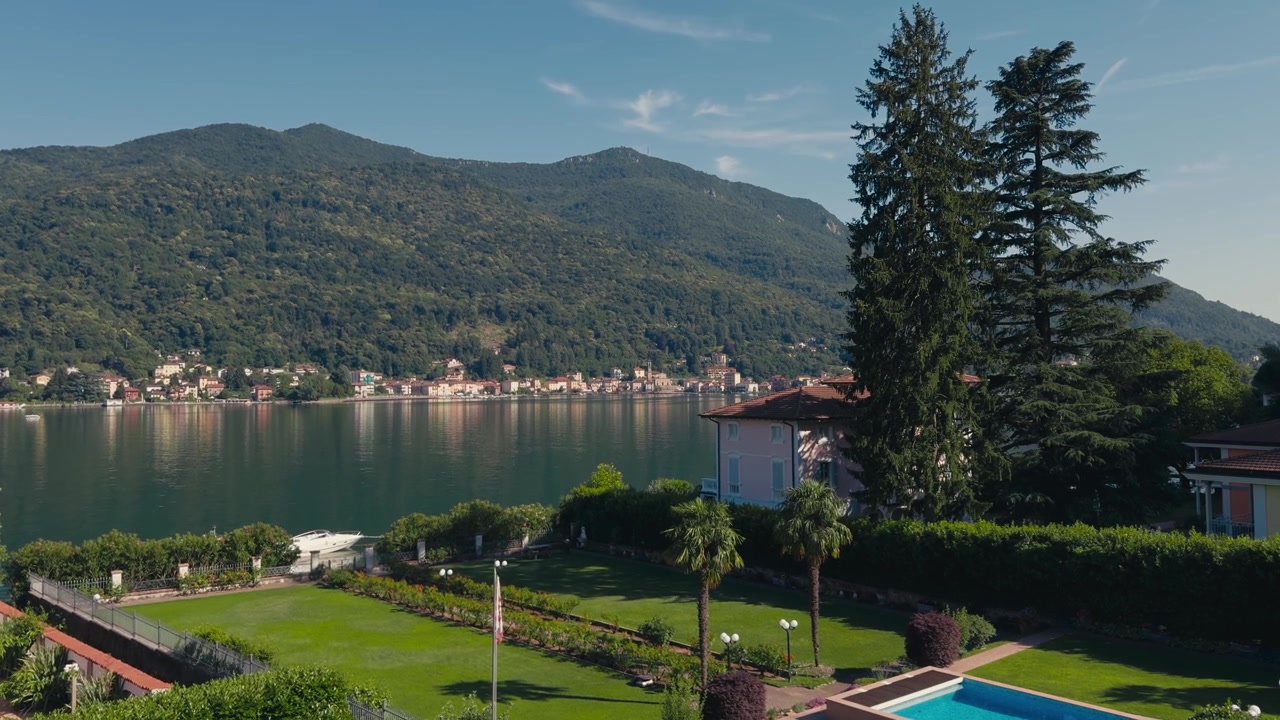 Vista statica dal giardino di un'elegante casa sul lago di Lugano con un prato curato, alberi maturi, una piscina, acqua calma e lussureggianti montagne verdi sotto un cielo estivo sereno.
