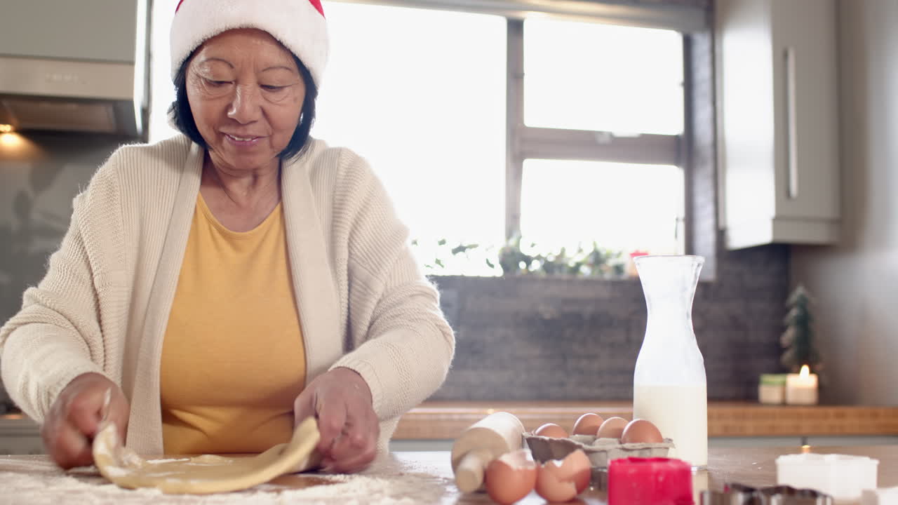 Senior Asian woman in Santa hat baking cookies at home, enjoying Christmas preparations