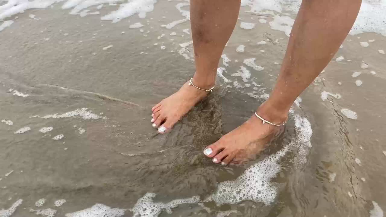 파도가 해변에 서 있는 발목걸이를 가진 소녀의 다리에 부 ⁇ 히고