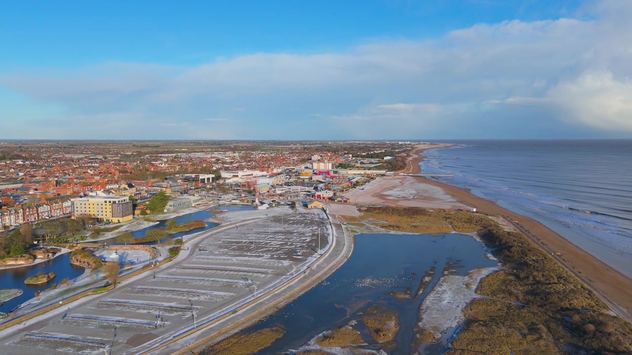 Prospettiva drone sopra una città costiera e un porto. Cielo azzurro brillante con nuvole sparse. L'area del molo presenta barche e acque calme. Il mare poco profondo incontra una spiaggia sabbiosa con dune coperte di alghe.