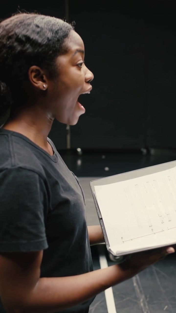 Vertical video: Singing woman following cue, rehearsing on black stage, holding music binder