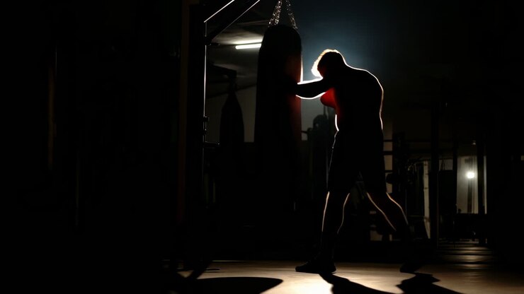 Boxeador en silueta entrenando en gimnasio, sesión nocturna con saco pesado, la iluminación de contorno resalta el perfil, guantes.