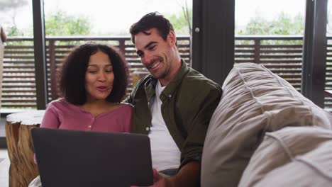 pareja de raza mixta sonriente usando computadora portátil juntos en el sofá en la casa de vacaciones