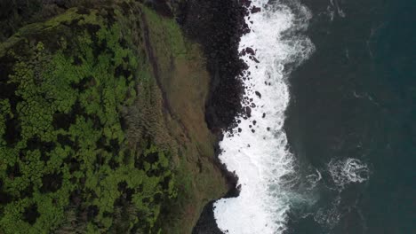 Top-down-view-of-waves-crashing-on-volcanic-island-bluff-with-lush-green-landscape-on-the-Azores-Islands