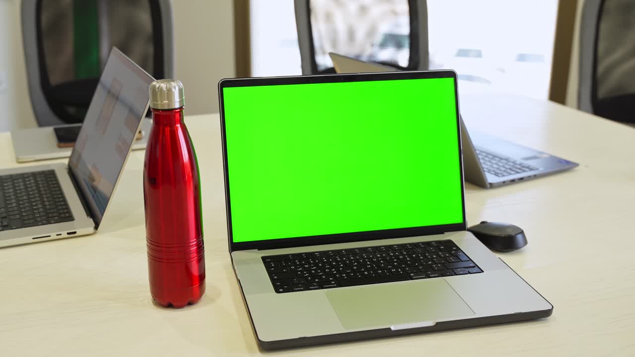Laptop with green screen in office on wooden desk with red bottle and mouse, ideal for VFX, tech scenes, presentations, or tutorials.