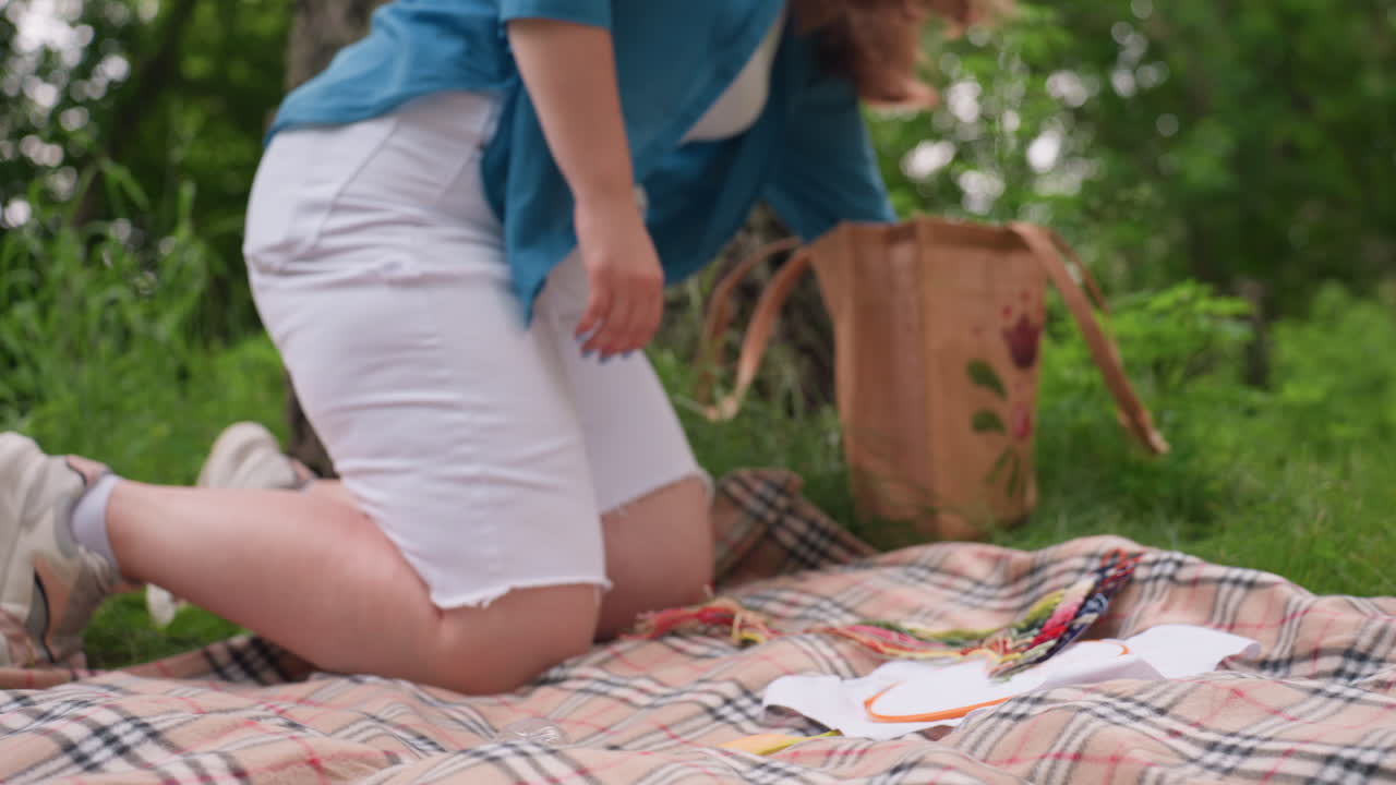 Close up of lady kneeling on patterned blanket taking water bottle, banana, and scissor from fabric bag, arranging them beside embroidery material in outdoor environment surrounded by green grass