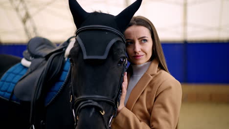 good-looking dark-haired lady standing close to a black horse. woman touching her cheek to horse's head. manege at backdrop in blur.