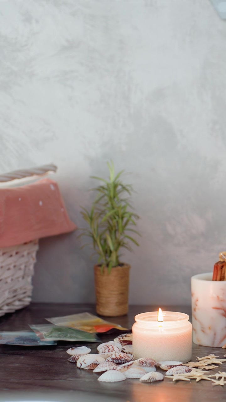 Bougie allumée sur une surface en bois entourée de coquillages, d'étoiles de mer et de petites plantes en pot sous une lumière naturelle douce. La scène présente une lumière de bougie chaude projetant de douces ombres sur les objets avec calme.