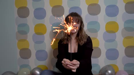 young woman sitting with a sparkler against colorful background, surrounded by balloons on the ground