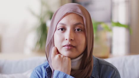 Videoporträt-Einer-Gemischtrassigen-Frau-In-Braunem-Hijab,-Die-Zu-Hause-Im-Wohnzimmer-In-Die-Kamera-Lächelt