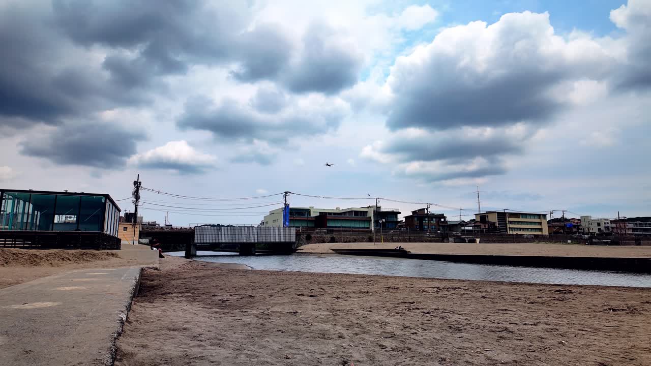 Wide static shot of a quiet sandy beach with canal and low-rise buildings, under a sky filled with dramatic clouds and two planes flying across the background in Kamakura Japan