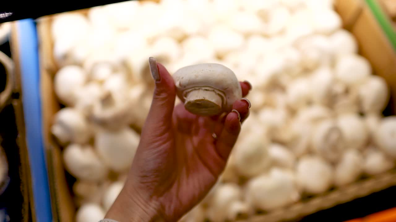 A hand examines white button mushrooms in a well-lit supermarket setting, highlighting freshness and quality selection