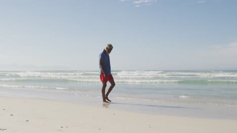 hombre afroamericano mayor caminando y tocando el agua en una playa soleada