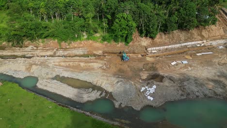 aerial orbit of heavy machinery working along riverbank and bridge structure in virac with water and rocks below forest edge