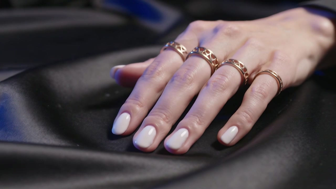 Placing woman's right hand making small shifts on black satin, revealing gold rings and white nails