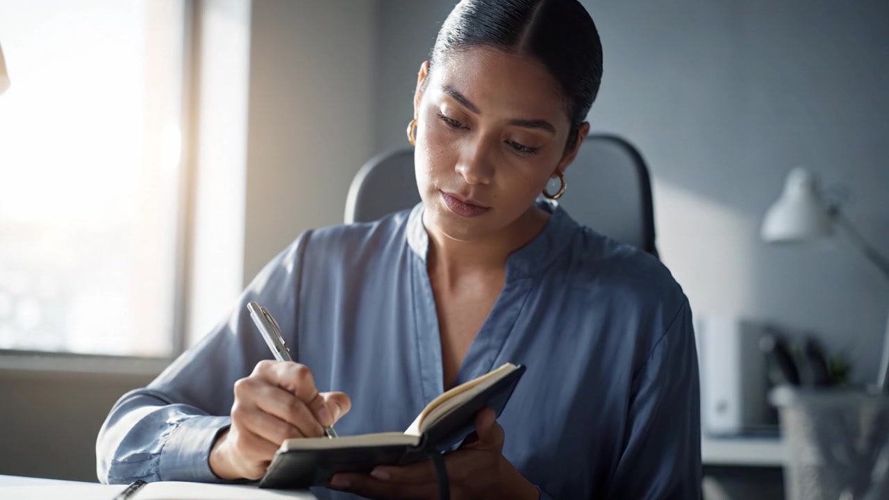 Focused individual jotting down notes in a notebook while seated at a desk in an office, emphasizing concentration and productivity in a light-filled workspace.