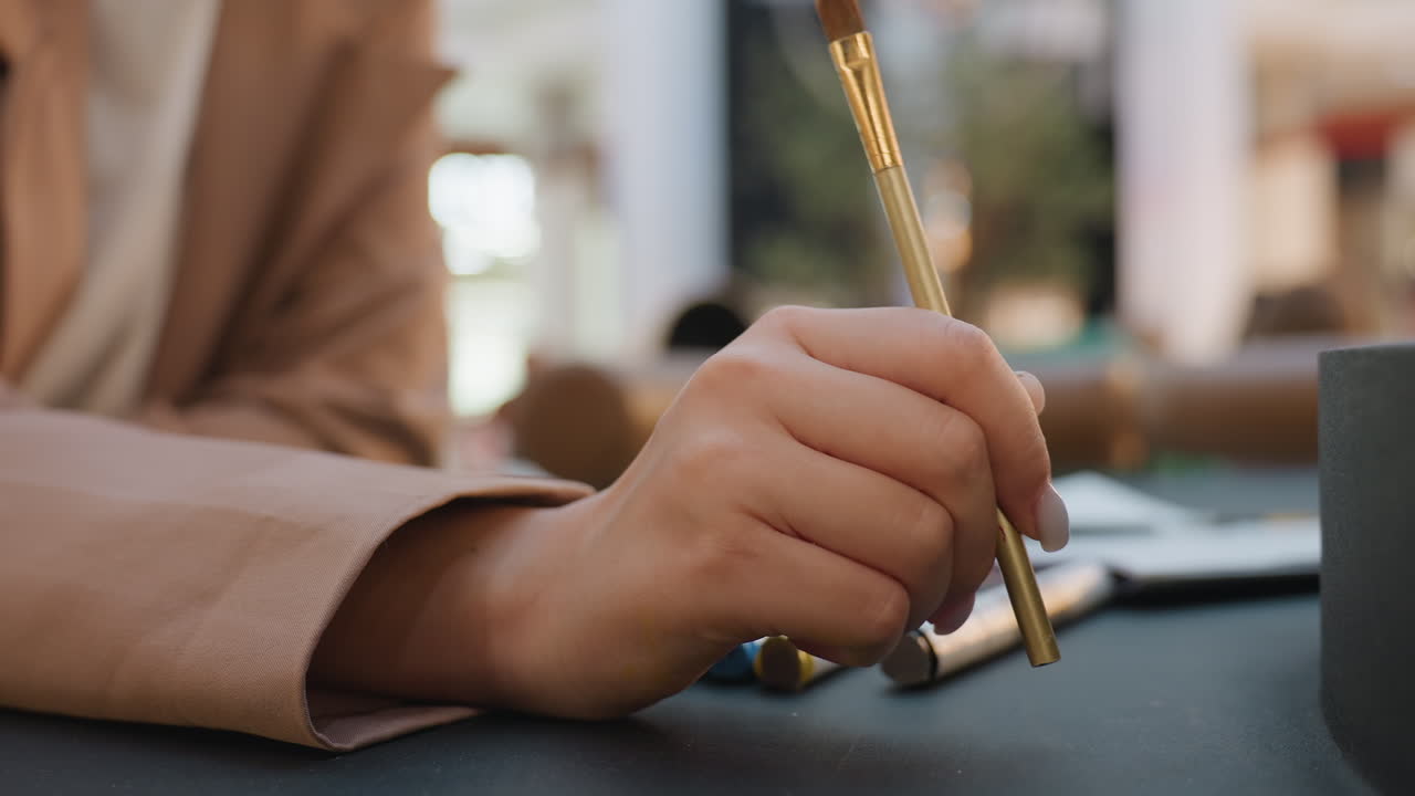 테이블 근처에서 금색 브러시를 들고 있는 손의 클로즈업, 빛을 받는 금속 손잡이, 천천히 균형 잡힌 움직임으로 브러시의 강모를 테스트, 세련된 우아함, 고급 카페에서 차분하고 신중한 제스처