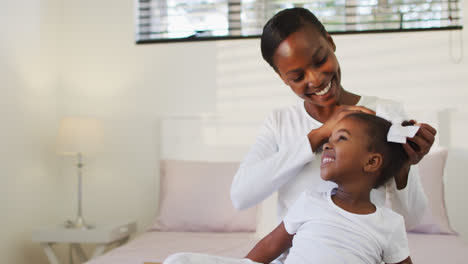 Feliz-Madre-E-Hija-Afroamericana-Sentadas-En-La-Cama,-Madre-Poniendo-Lazo-Para-El-Pelo