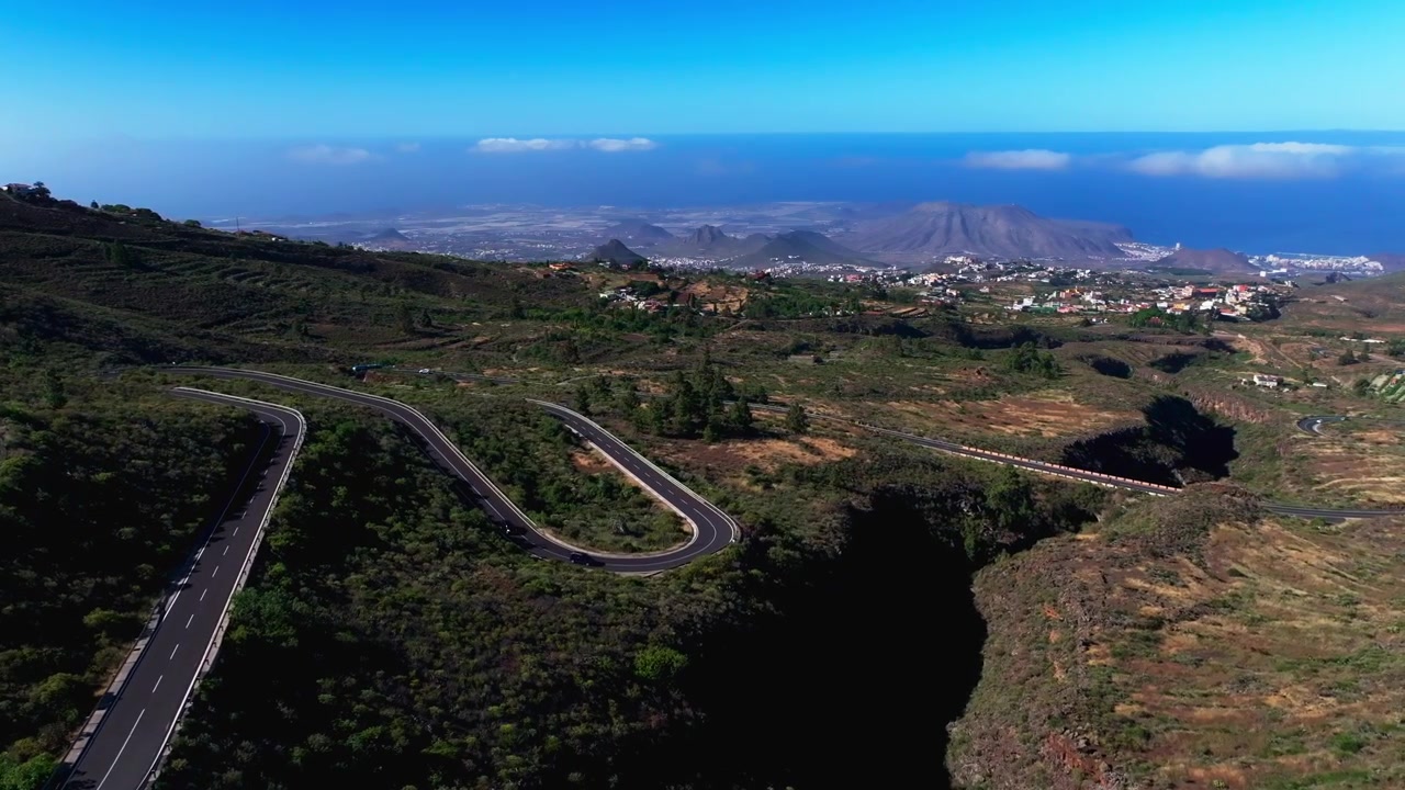 Vista aerea di una strada di montagna tortuosa attraverso terreno vulcanico con una città costiera in lontananza.