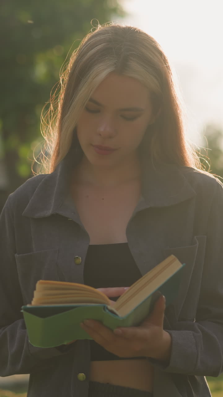 dama en ropa gris de pie al aire libre hojeando las páginas de un libro, con la luz del sol suave iluminando a su alrededor y un fondo borroso de árboles y un edificio