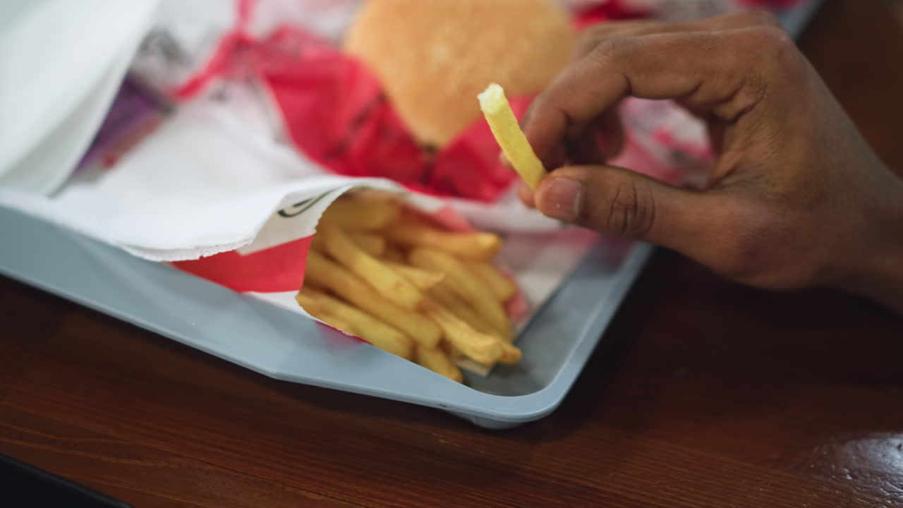 Close view of hand picking french fries from paper tray, golden crispy pieces with slight grease sheen, fingers grasping one fry, soft background blur emphasizing texture and appetite