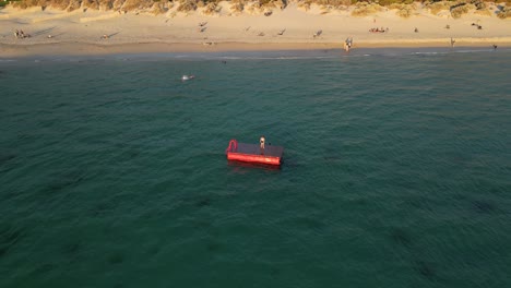 Aerial-top-down-shot-of-two-friends-on-floating-platform-on-ocean-having-fun-during-golden-sunset---South-Beach-of-Fremantle,Western-Australia---Drone-orbiting-flight