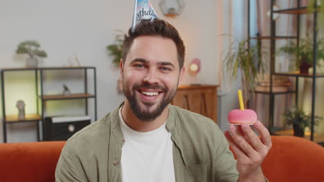 happy young man wears birthday hat makes wish congratulating blowing burning candle on cake at home