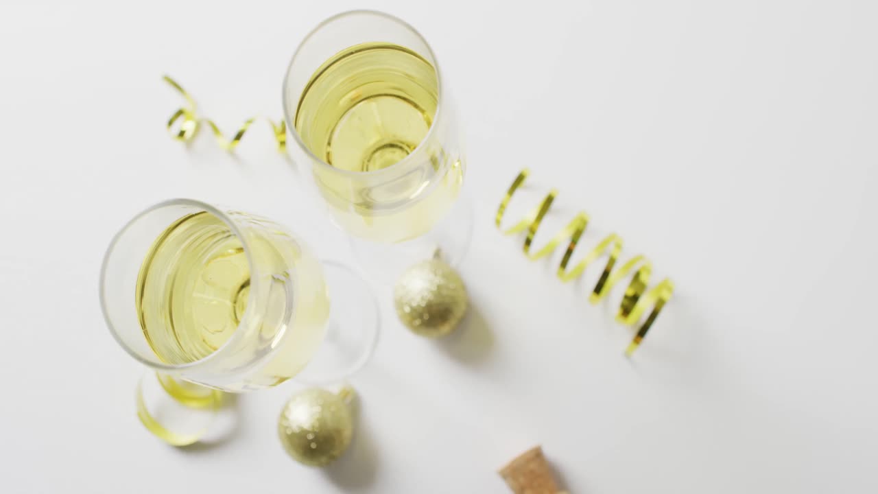 Video dall'alto di due bicchieri di champagne pieni con stelle filanti dorate e palline decorative, su sfondo bianco.