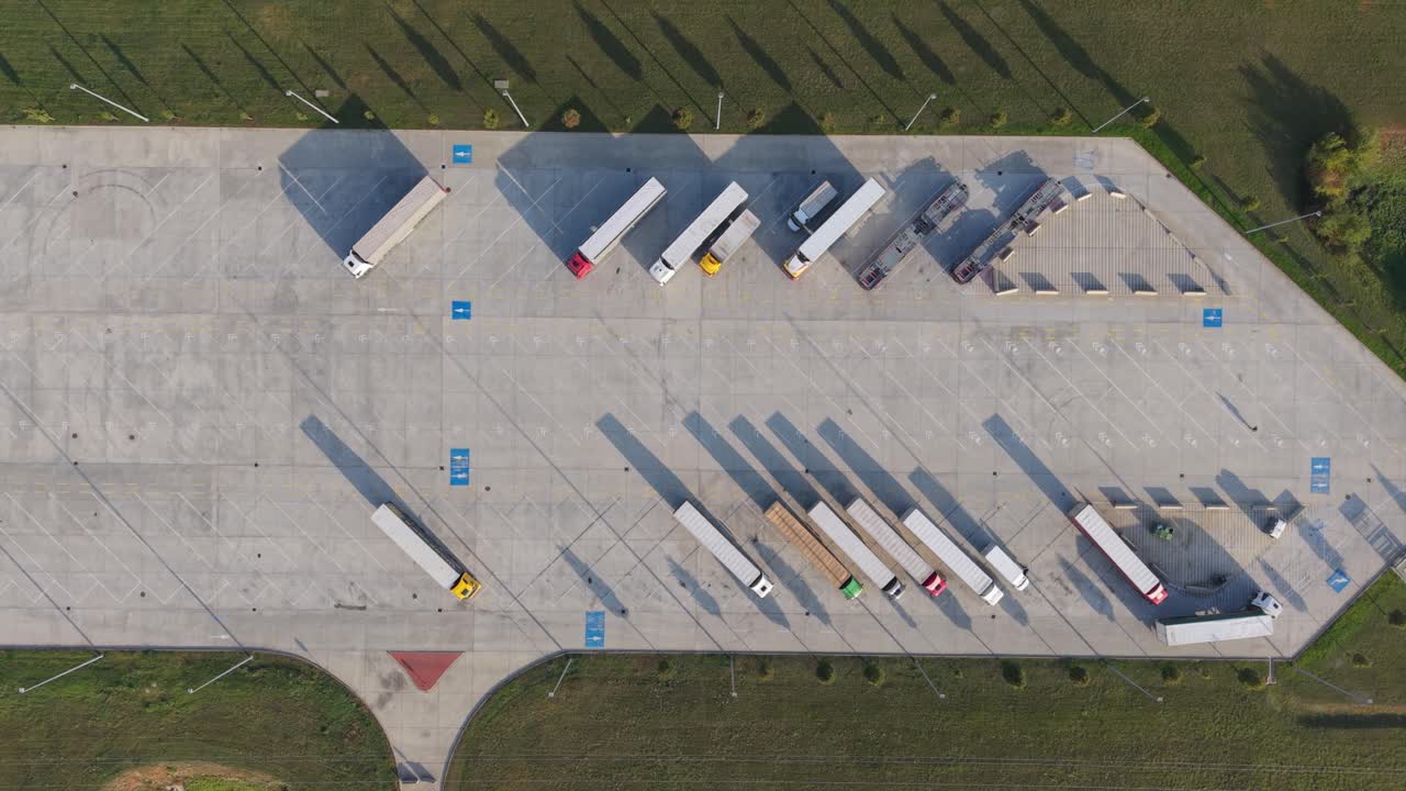 A top-down aerial view of a large truck parking lot showing multiple semi-trailers parked in designated spaces, while one truck leaving the area