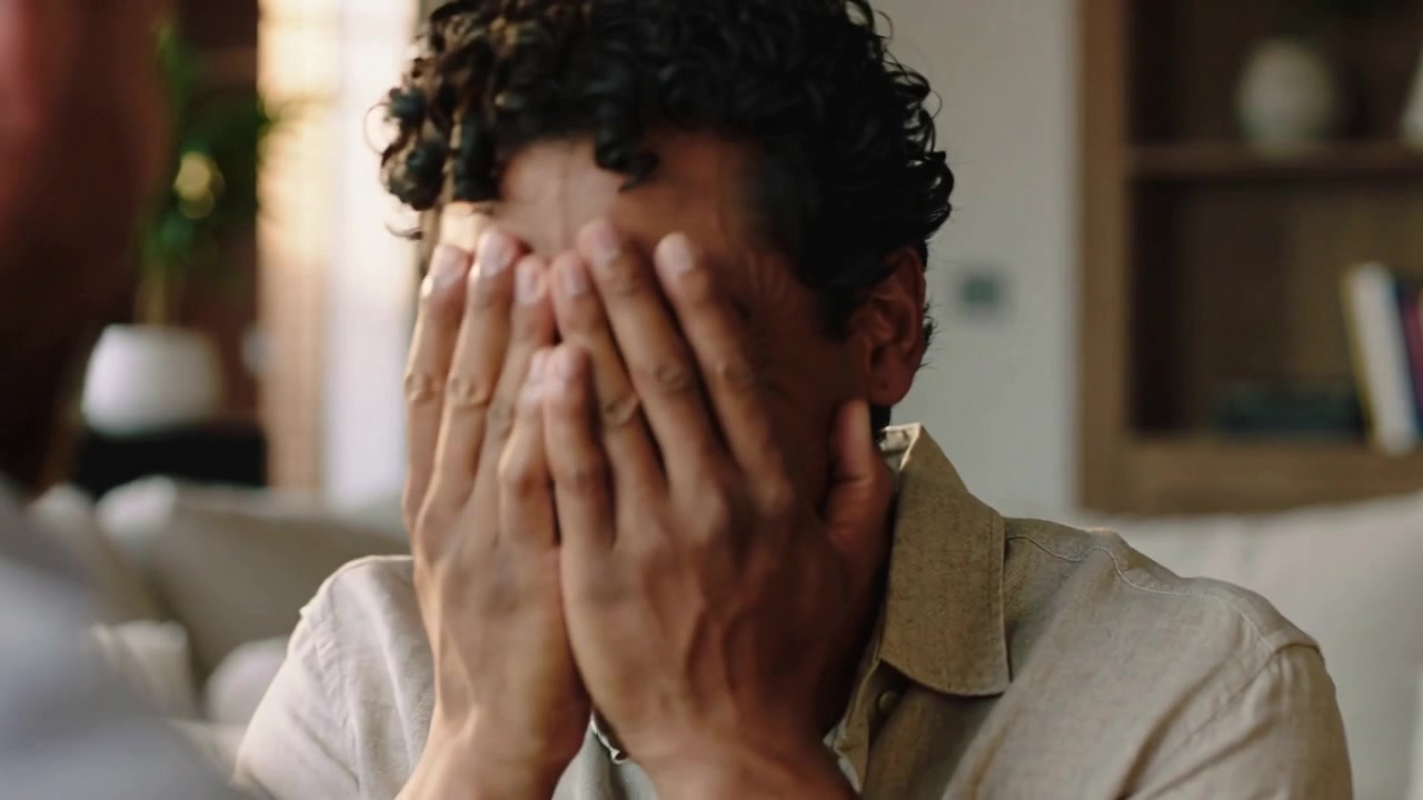 Laughing man covering face while reacting to offscreen speaker in living room with sofa, shirt