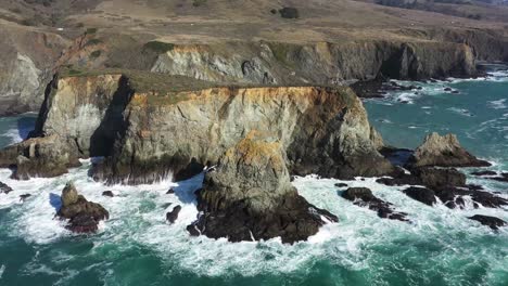 Aerial-view-of-ocean-waves-crashing-against-large-cliff-rocks-with-slow-pan-up-and-out