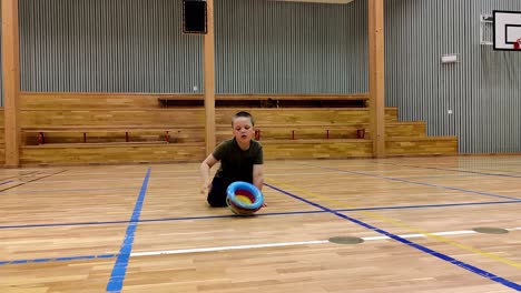 Boy-with-autism-and-sensory-disorders-sitting-on-gymnasium-floor-and-spinning-a-colorful-plastic-toy-in-circles---Static-handheld-clip-with-boy-in-center