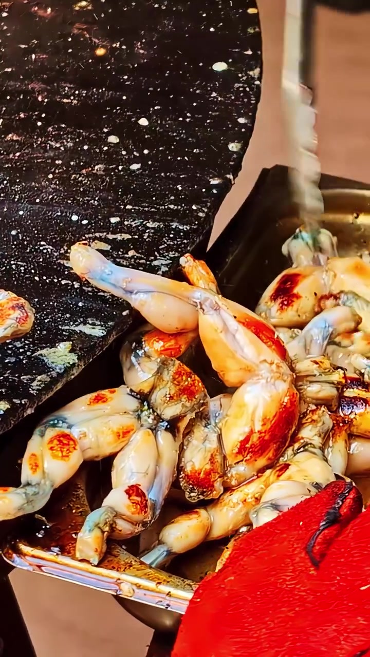 Metal tongs collecting freshly grilled frog legs from hot black plancha into serving container at outdoor street food festival, pan right vertical video