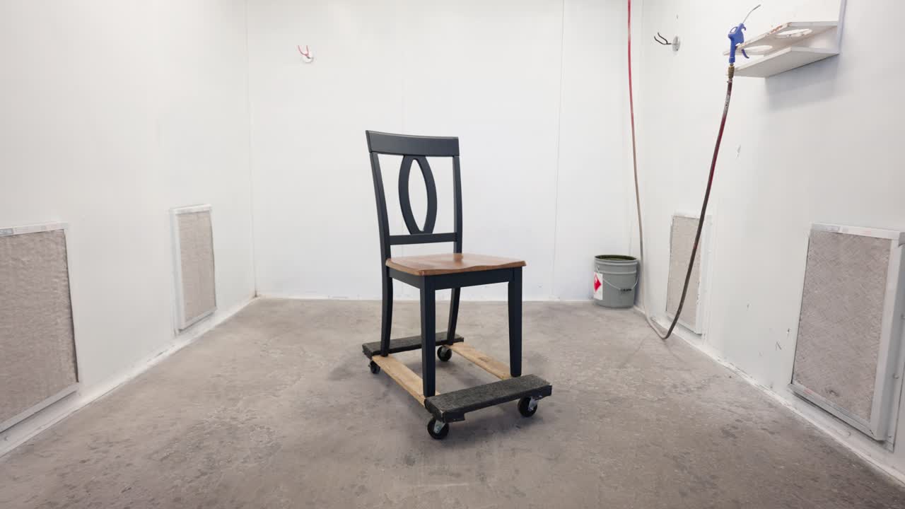 Camera dollies out from grey wood dining chair in spray booth, factory interior