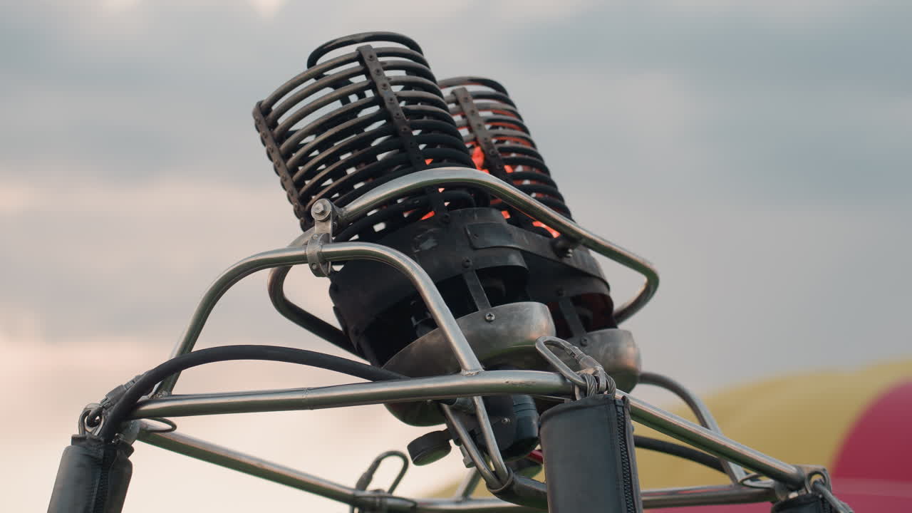 metal burner of hot air balloon glowing bright flame beneath steel frame controls while pilot adjusts handle in grassy field at dusk preparing flight over tranquil rural landscape