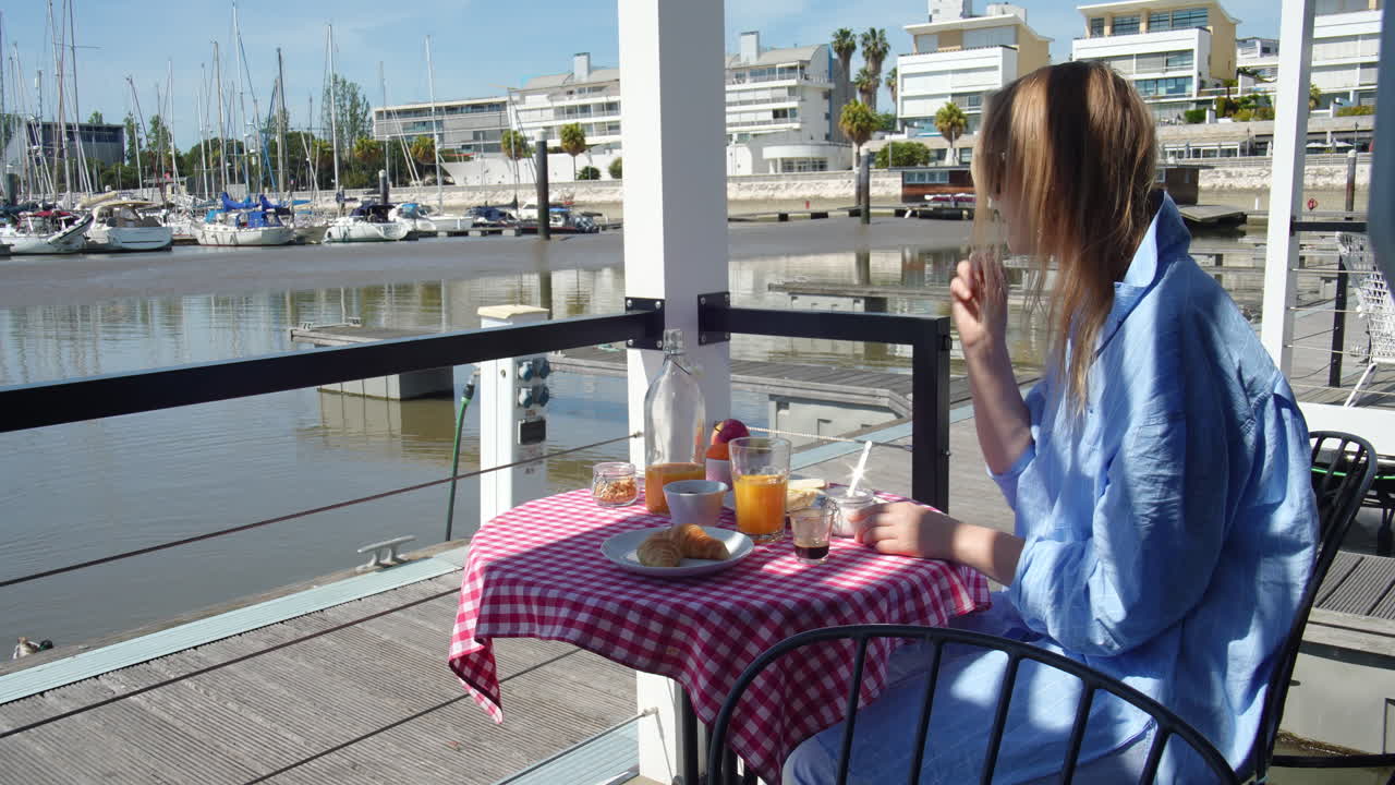 Young woman relaxing in blue bathrobe, enjoying morning breakfast at waterfront marina, sipping orange juice and savoring pastry with scenic harbor and boats surrounding her