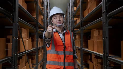 ingeniero masculino asiático con casco de seguridad de pie en el almacén con estantes llenos de mercancías de entrega. sonriendo y tocando su pecho luego apuntando a usted en el almacenamiento