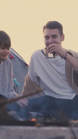 joyful young tourists drink tea resting at burning bonfire in camp with tent in warm autumn evening closeup slow motion