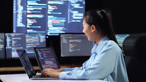 side view of asian female programmer looking at database on tablet and writing code by a laptop using multiple monitors showing database on desktops in the office