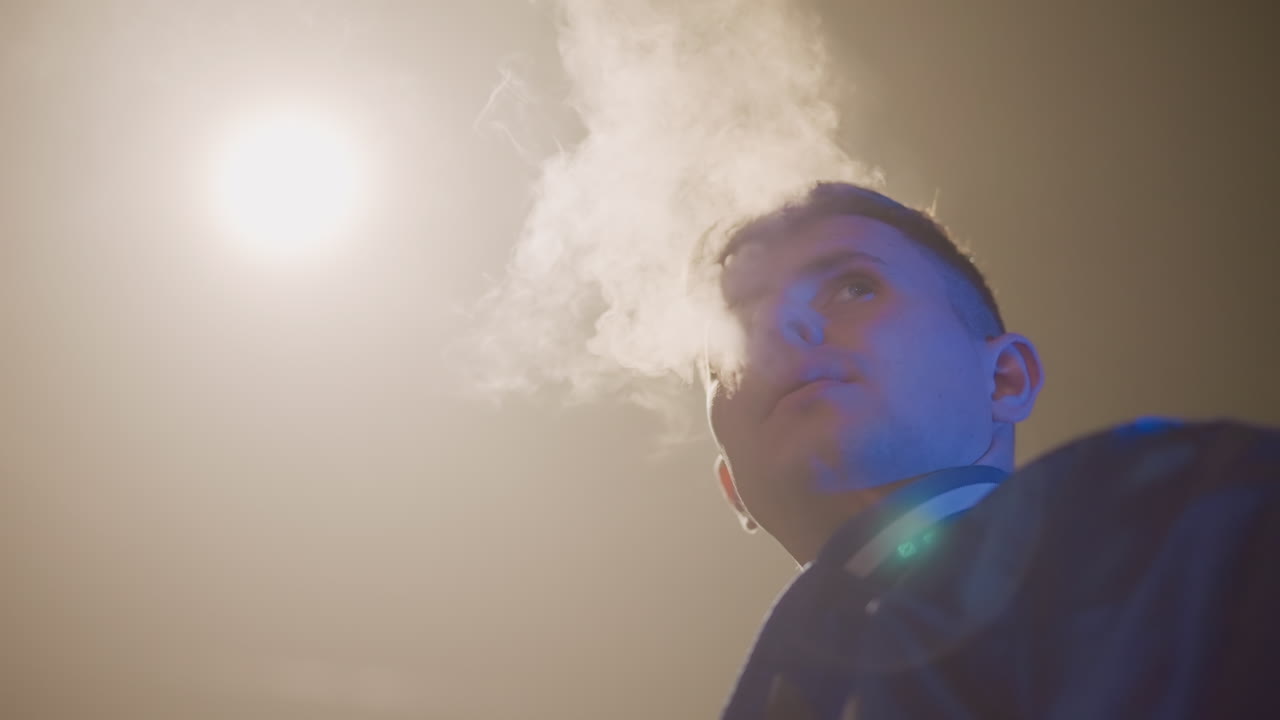 거리의 가로등 아래에서 전자 담배를 내뿜는 젊은 백인, 로우 앵글의 시네마틱 클로즈업, 손에 들고 있는 장치의 빛, 짙은 증기 기둥, 파란색 재킷, 렌즈 플레어, 거친 밤의 분위기, 운전 중 휴식