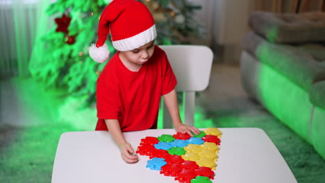 kid making xmas decorations. a young child wearing a santa hat assembles colorful shapes on a table while a festive tree glows nearby